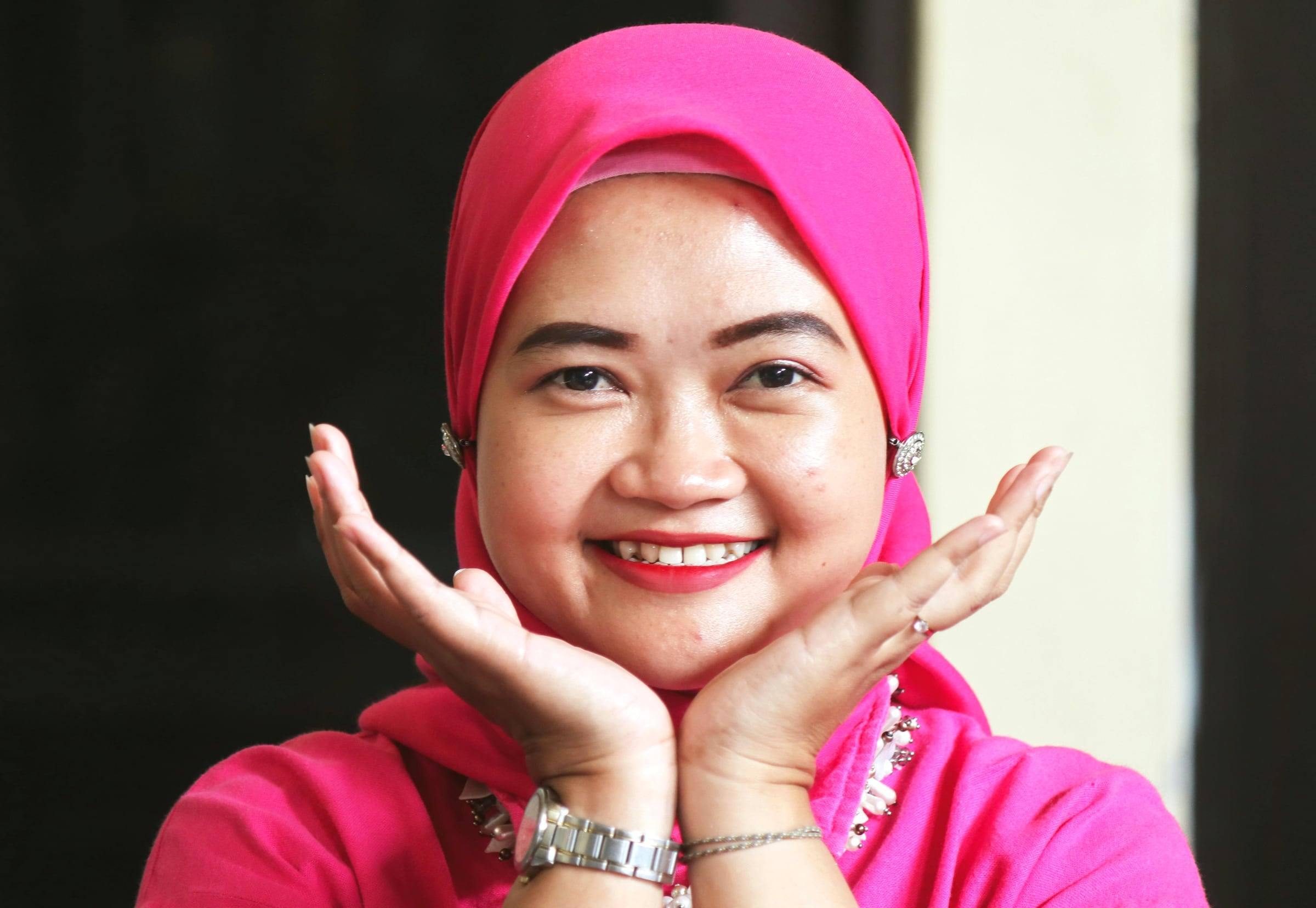 A woman in a pink headscarf smiling broadly, her hands framing her face.