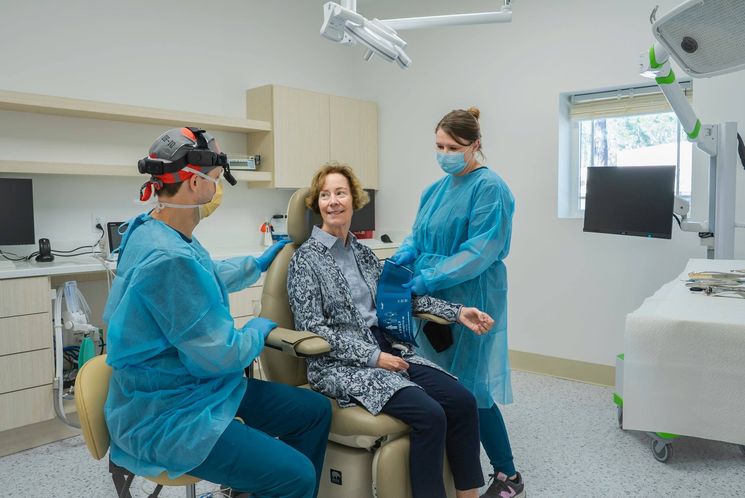 A smiling patient in a dental chair talking with two masked dental staff in a bright room.